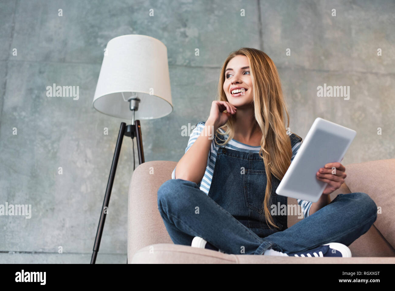 young smiling woman sitting on pink sofa in room with digital tablet in ...