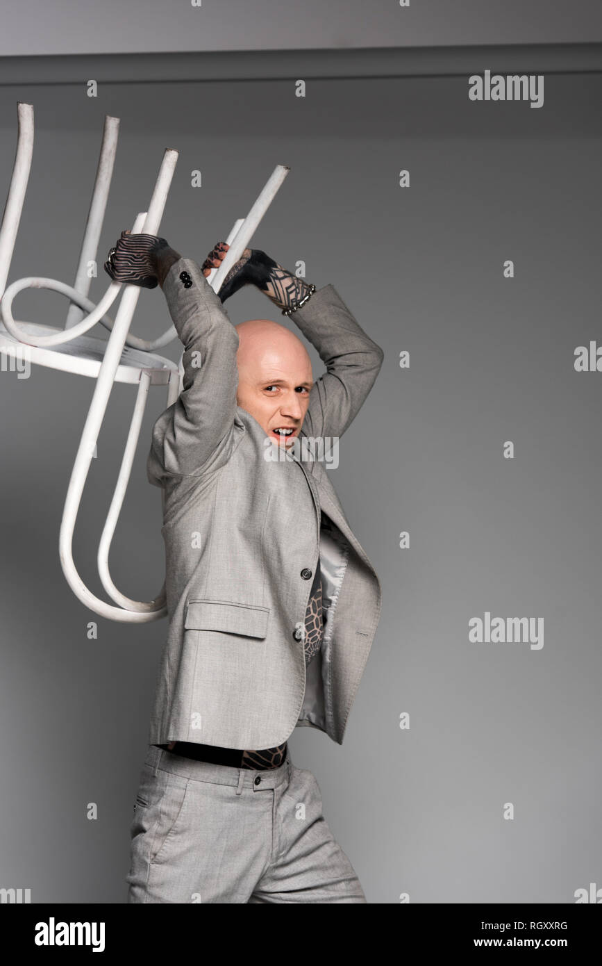 aggressive tattooed man in suit holding chair above head and looking at camera on grey Stock Photo