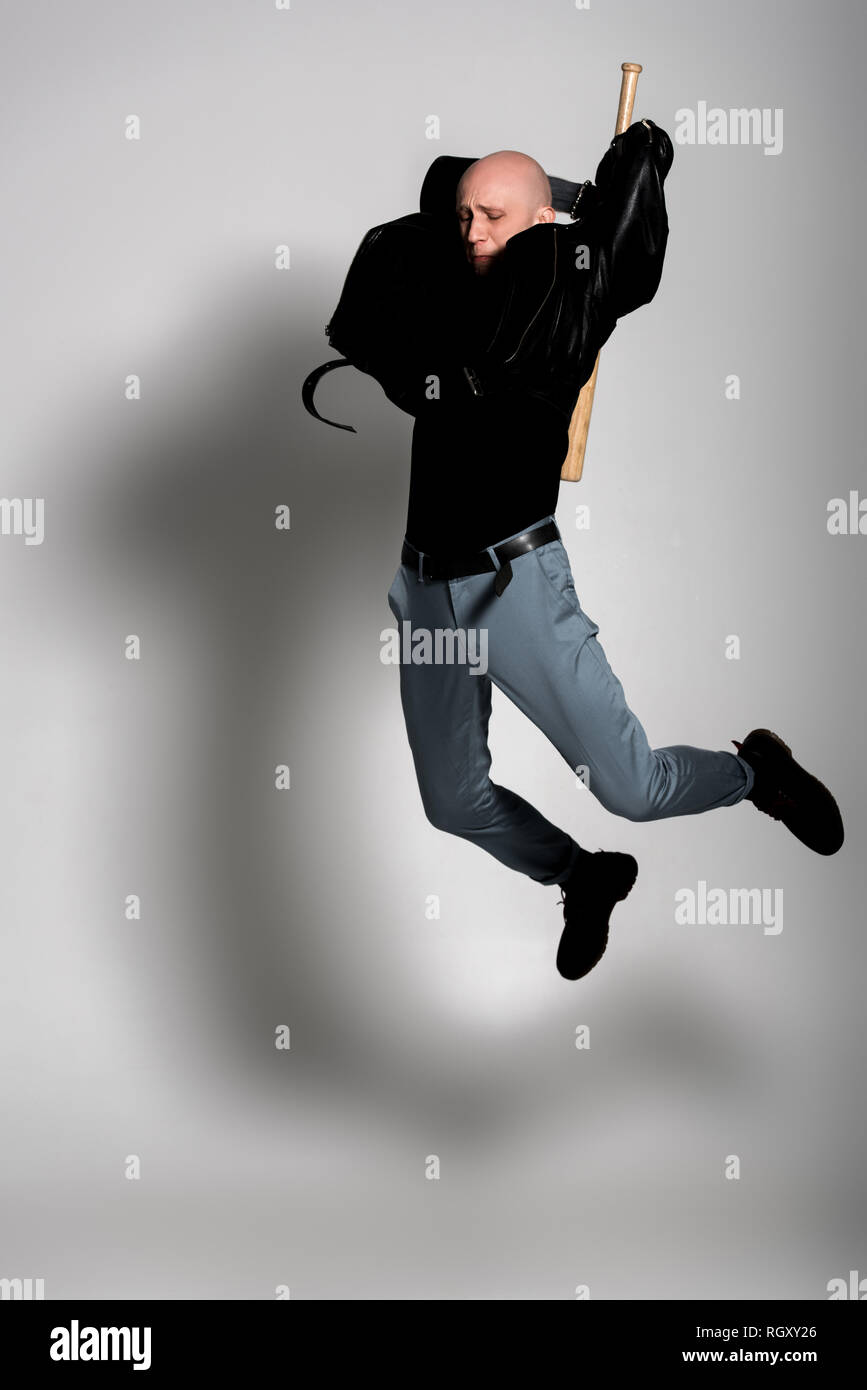 aggressive bald man in leather jacket holding baseball bat and jumping on grey Stock Photo