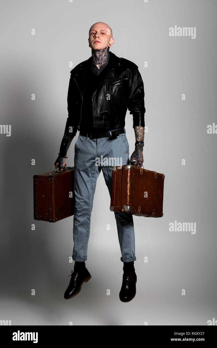 full length view of handsome tattooed man in leather jacket holding suitcases and jumping on grey Stock Photo