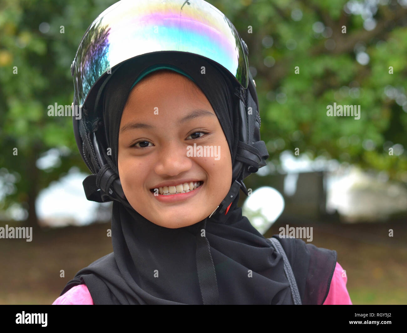 Smiling, pretty Muslim Malay girl wears a modern motorcycle crash helmet over her black traditional hijab. Stock Photo