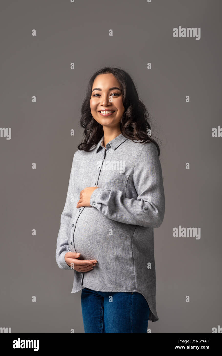 Smiling young pregnant asian woman holding tummy with hands isolated on grey Stock Photo
