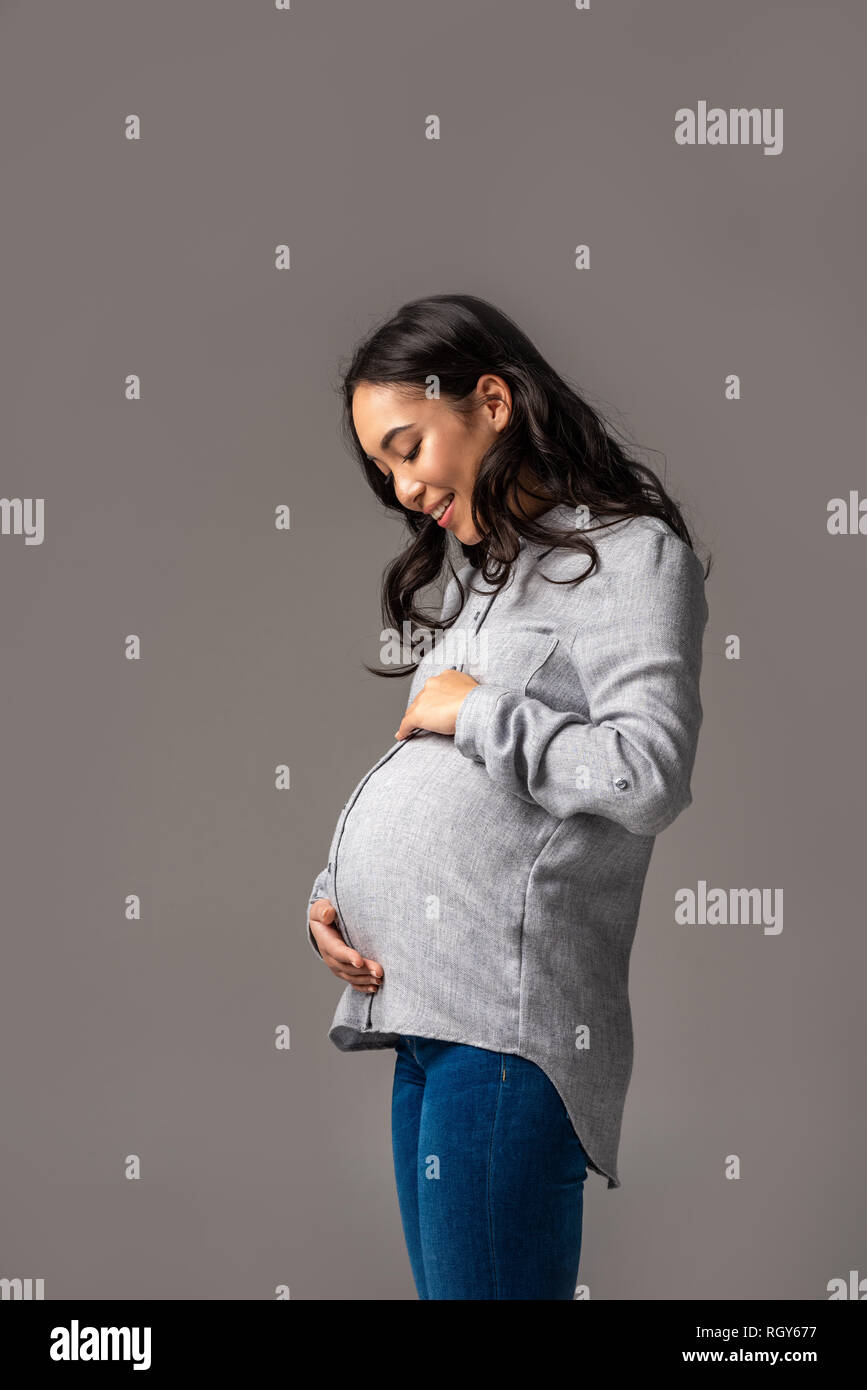 Happy pregnant asian woman looking at belly with smile isolated on grey Stock Photo