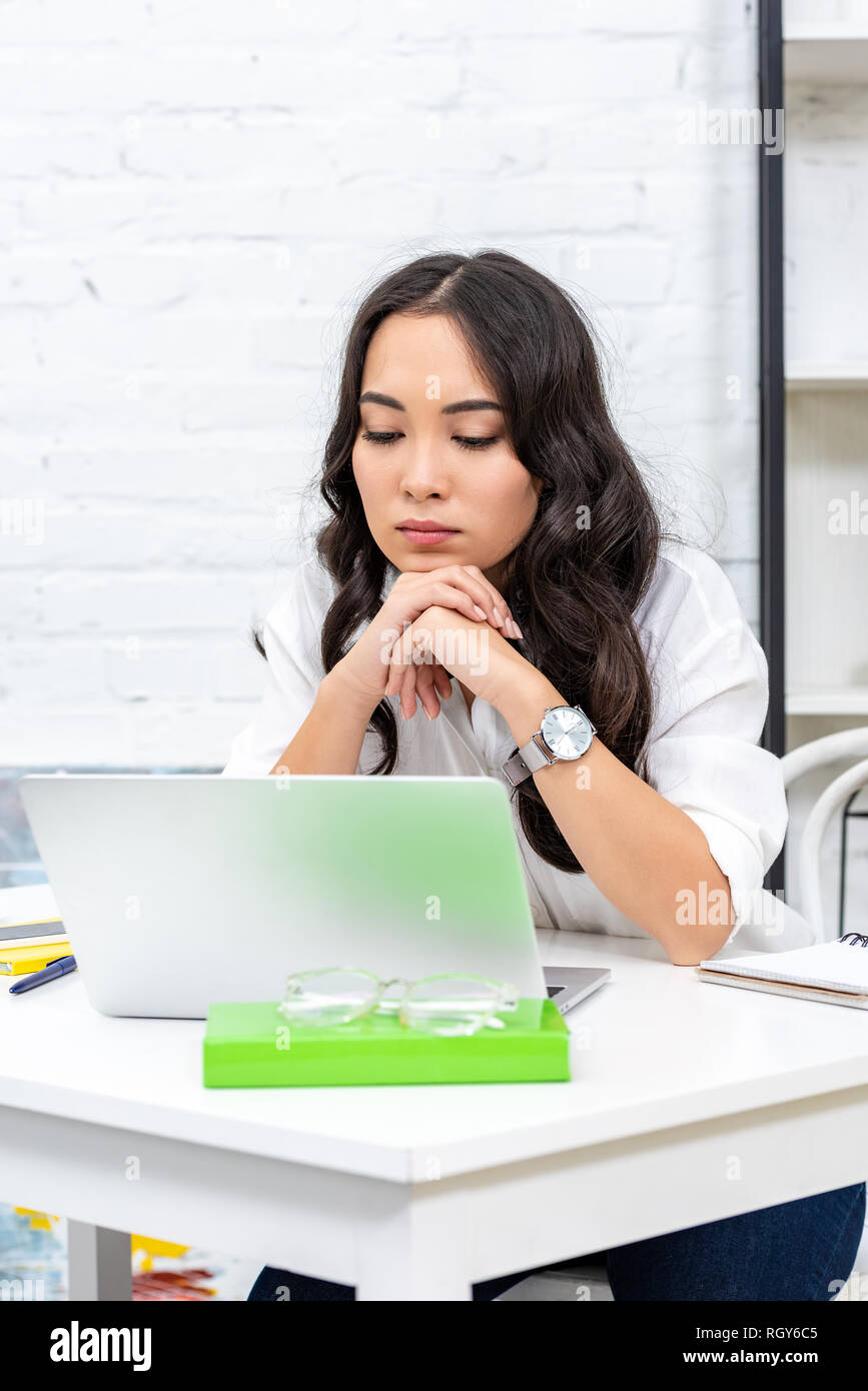 Tired asian freelancer looking at laptop screen and clutching hands under chin Stock Photo