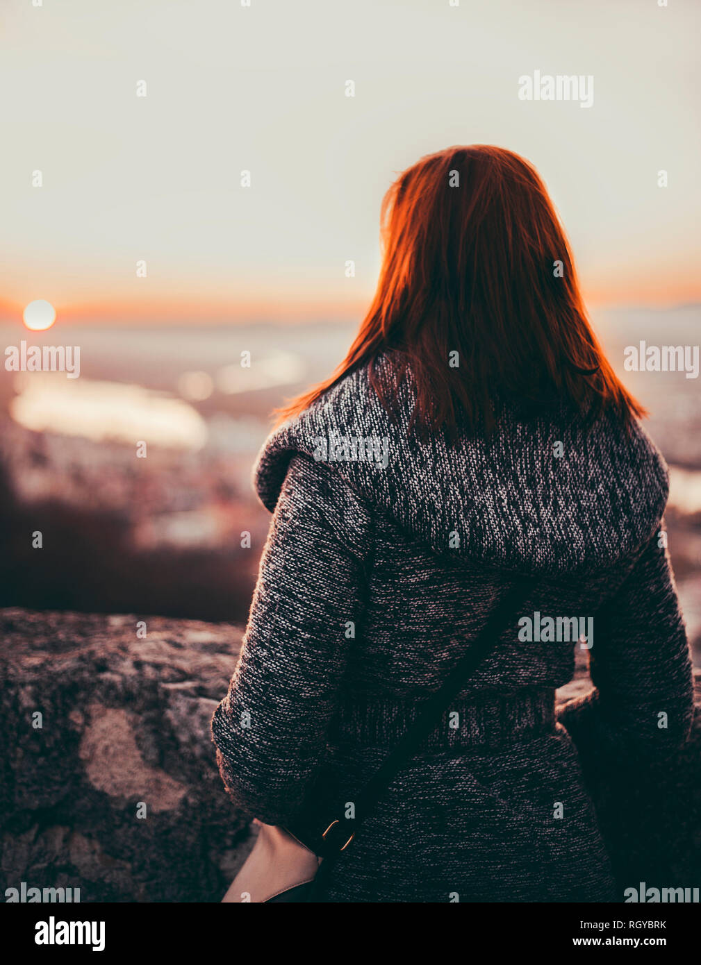 Photo of red haired woman in coat standing on the medieval walls over the city on sunset in autumn. Girl with hands in pockets looking on sunset scene Stock Photo