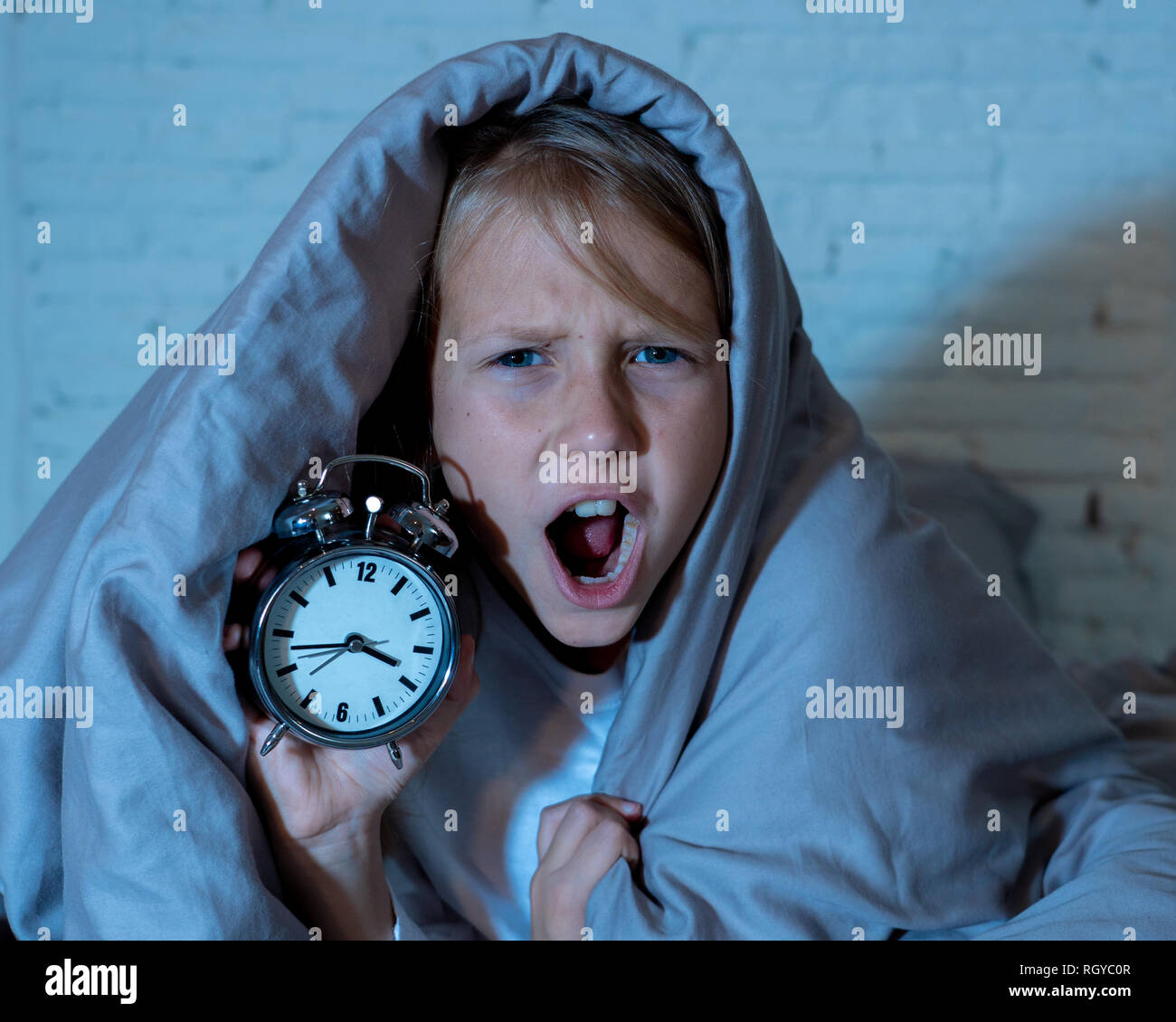 Cute sleepless little girl lying in bed showing alarm clock looking