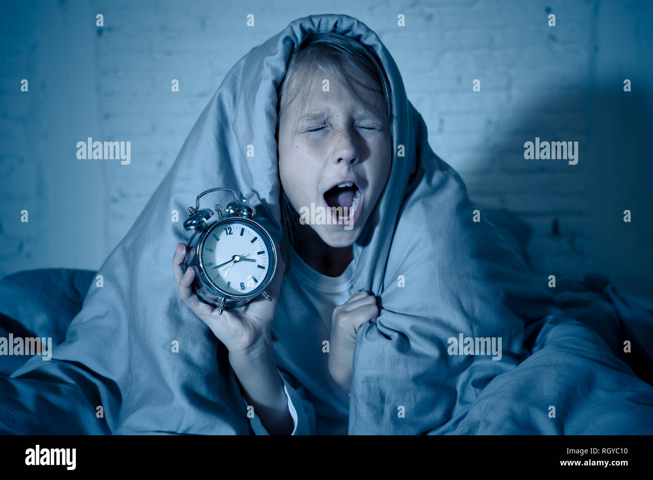 Cute sleepless little girl lying in bed showing alarm clock looking