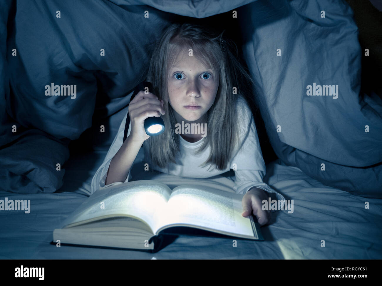 Beautiful caucasian girl lying in bed under the duvet holding a lantern reading a mystery book in the dark late at night looking frightened with a dra Stock Photo