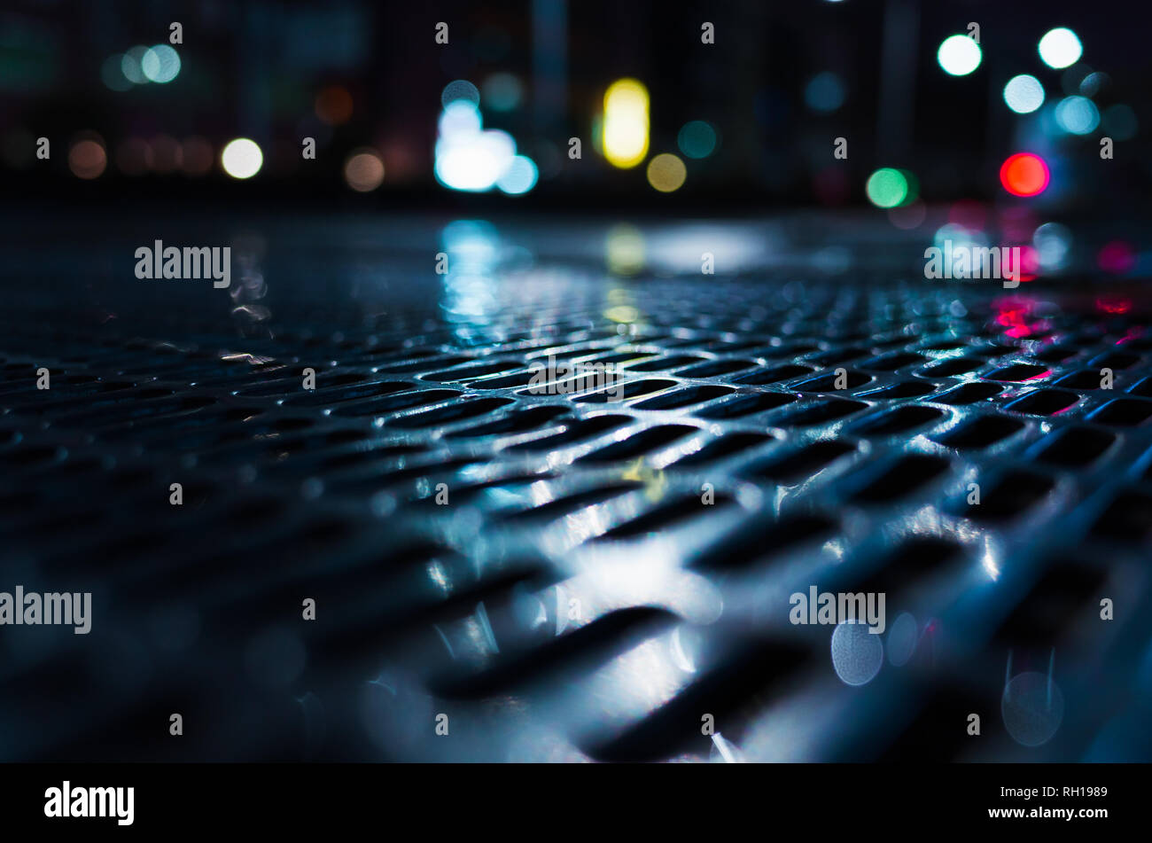 Abstract night city background with shiny wet street sewer grate on urban road, close-up photo with selective focus Stock Photo