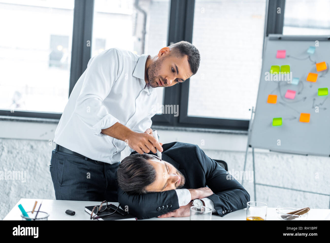 focused man drawing with marker on face of sleeping coworker in modern office Stock Photo