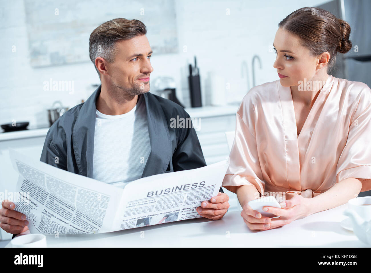 man in robe reading business newspaper while woman using smartphone during breakfast in kitchen Stock Photo