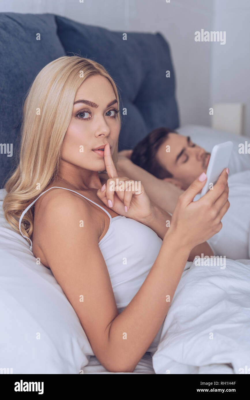 young woman gesturing for silence and looking at camera while boyfriend sleeping in bed, secret concept Stock Photo
