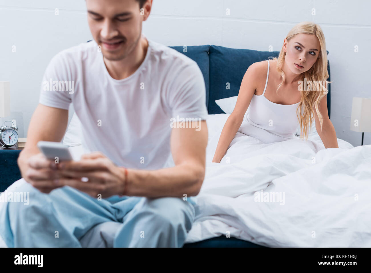 jealous young woman looking at boyfriend using smartphone on foreground in bedroom Stock Photo