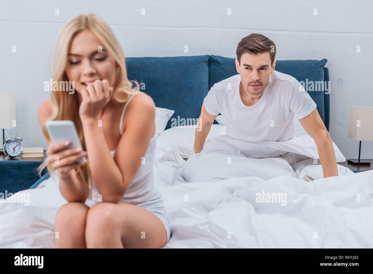 beautiful young woman using smartphone and jealous boyfriend peeking behind on bed Stock Photo