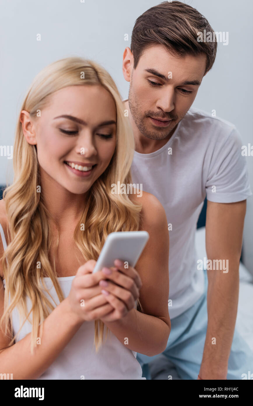 jealous young man looking at beautiful smiling wife using smartphone in bedroom Stock Photo