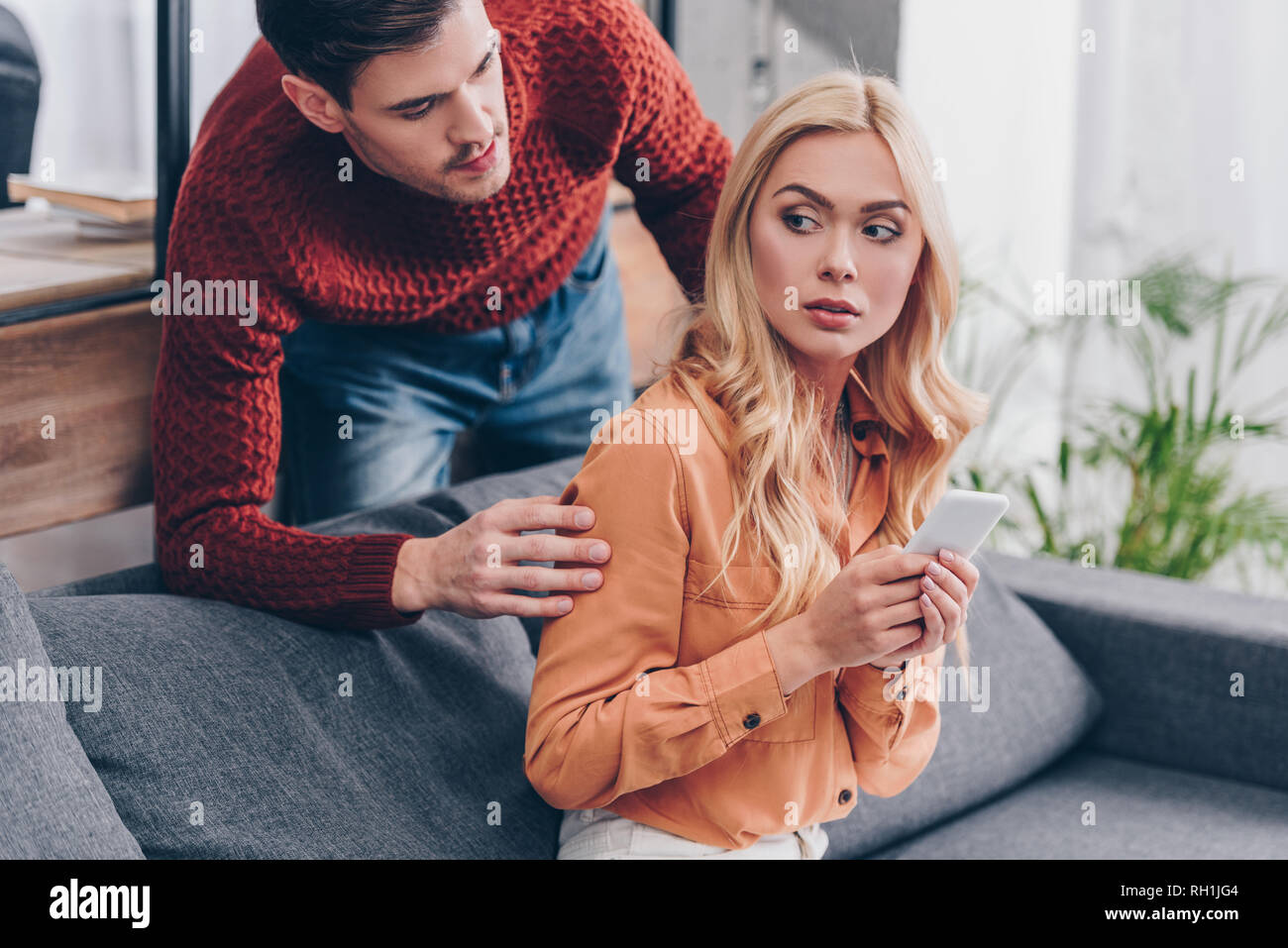 jealous husband looking at scared young woman using smartphone on couch, distrust concept Stock Photo