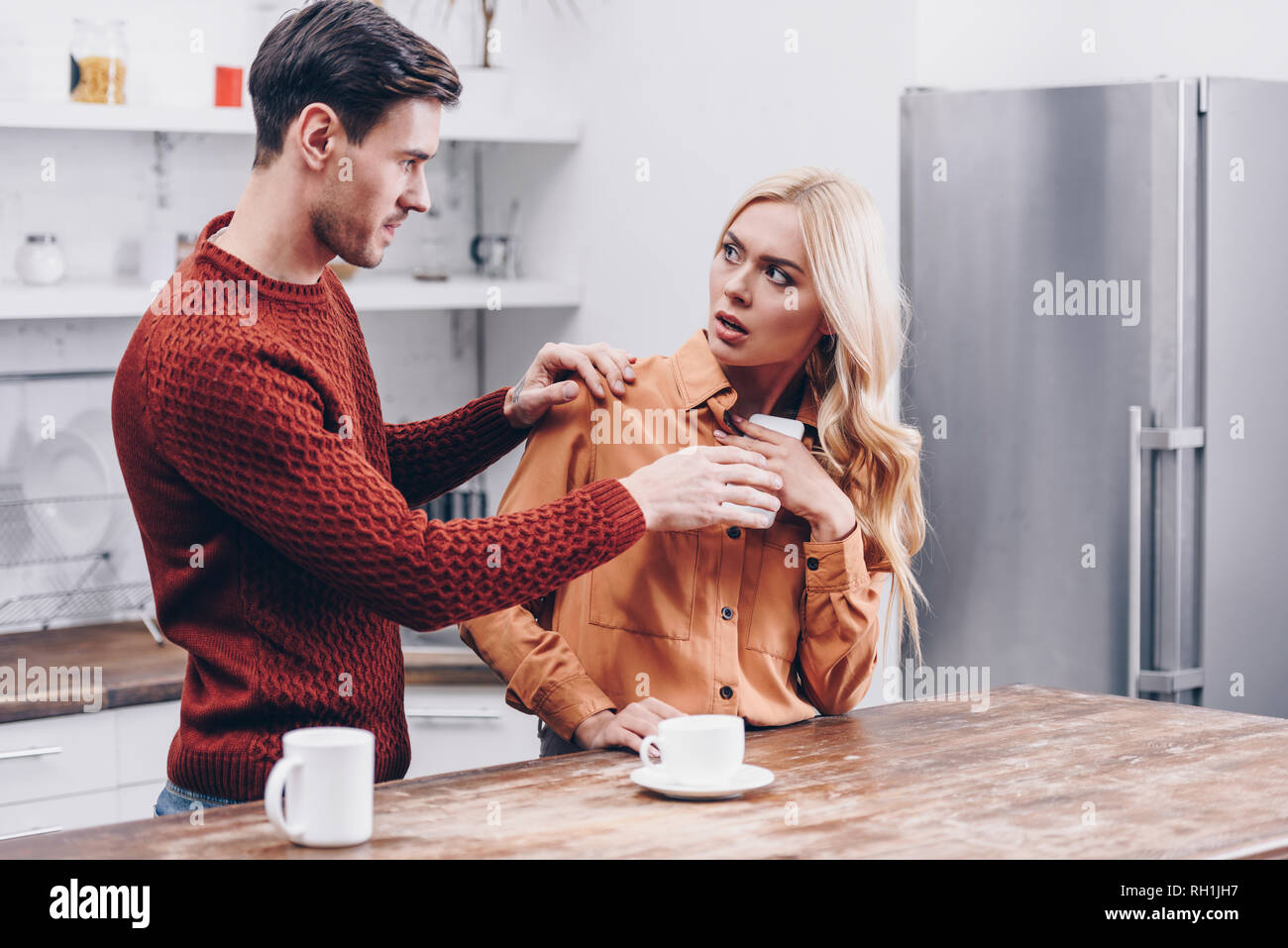 jealous young man looking at scared girlfriend using smartphone in kitchen Stock Photo