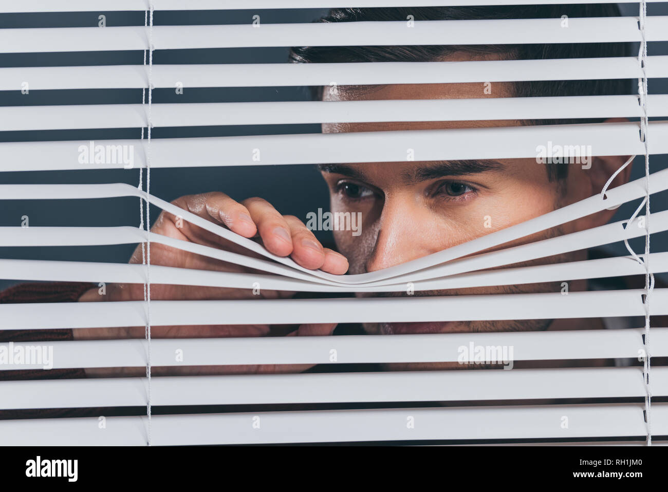 suspicious young man looking away and peeking through blinds Stock