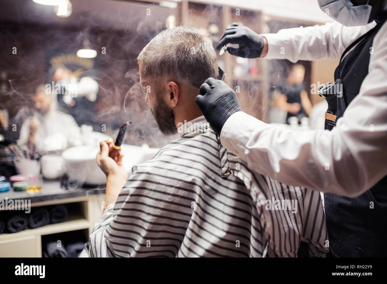 Handsome hipster man client visiting haidresser and hairstylist in barber shop, smoking a pipe. Stock Photo