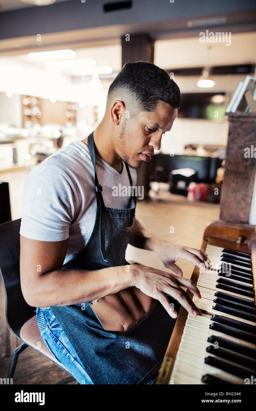 Young male hispanic haidresser and hairstylist sitting in barber shop, playing piano. Stock Photo