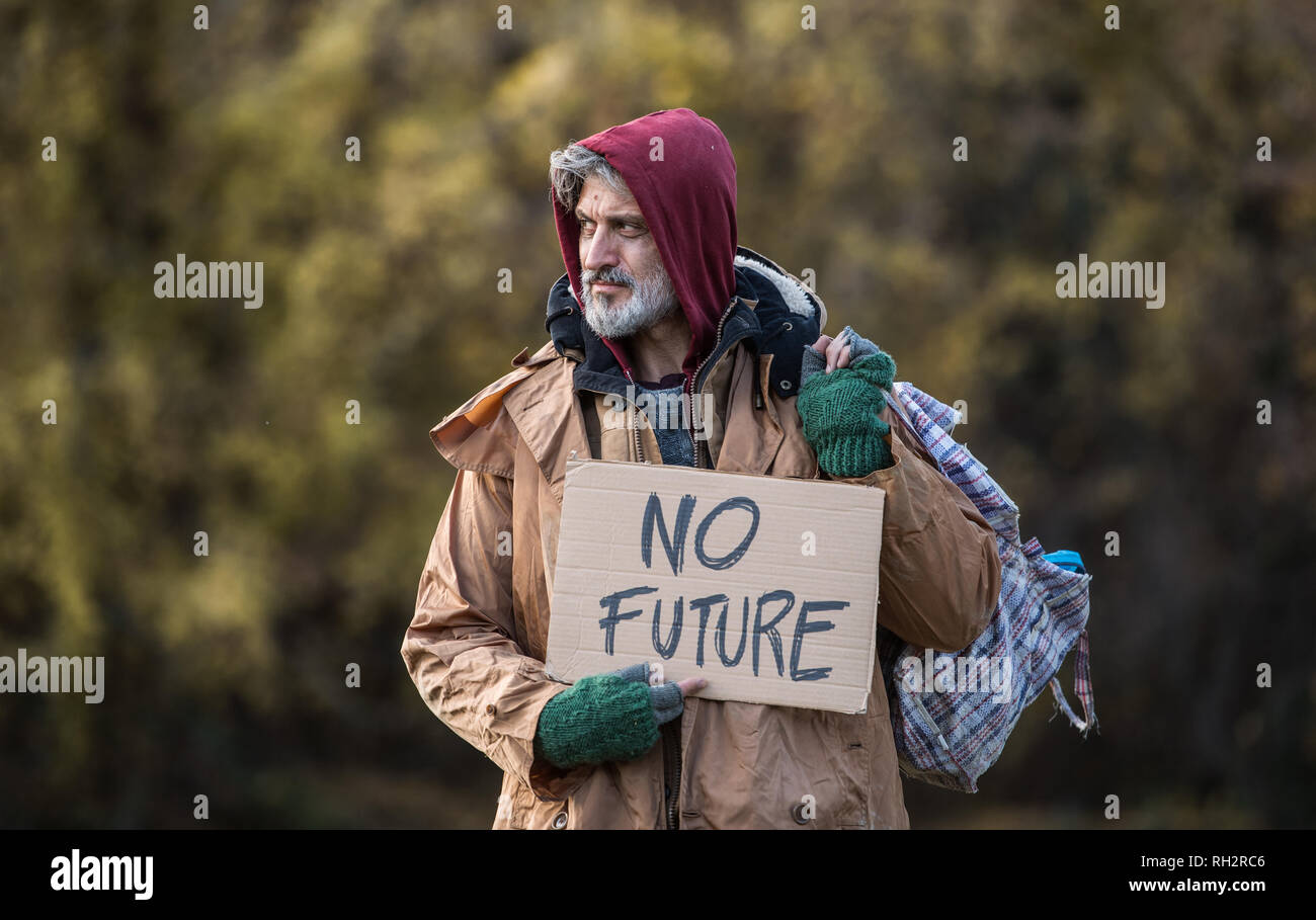 A portrait of homeless beggar man standing outdoors in park in autumn, holding bag and no future cardboard sign. Copy space. Stock Photo