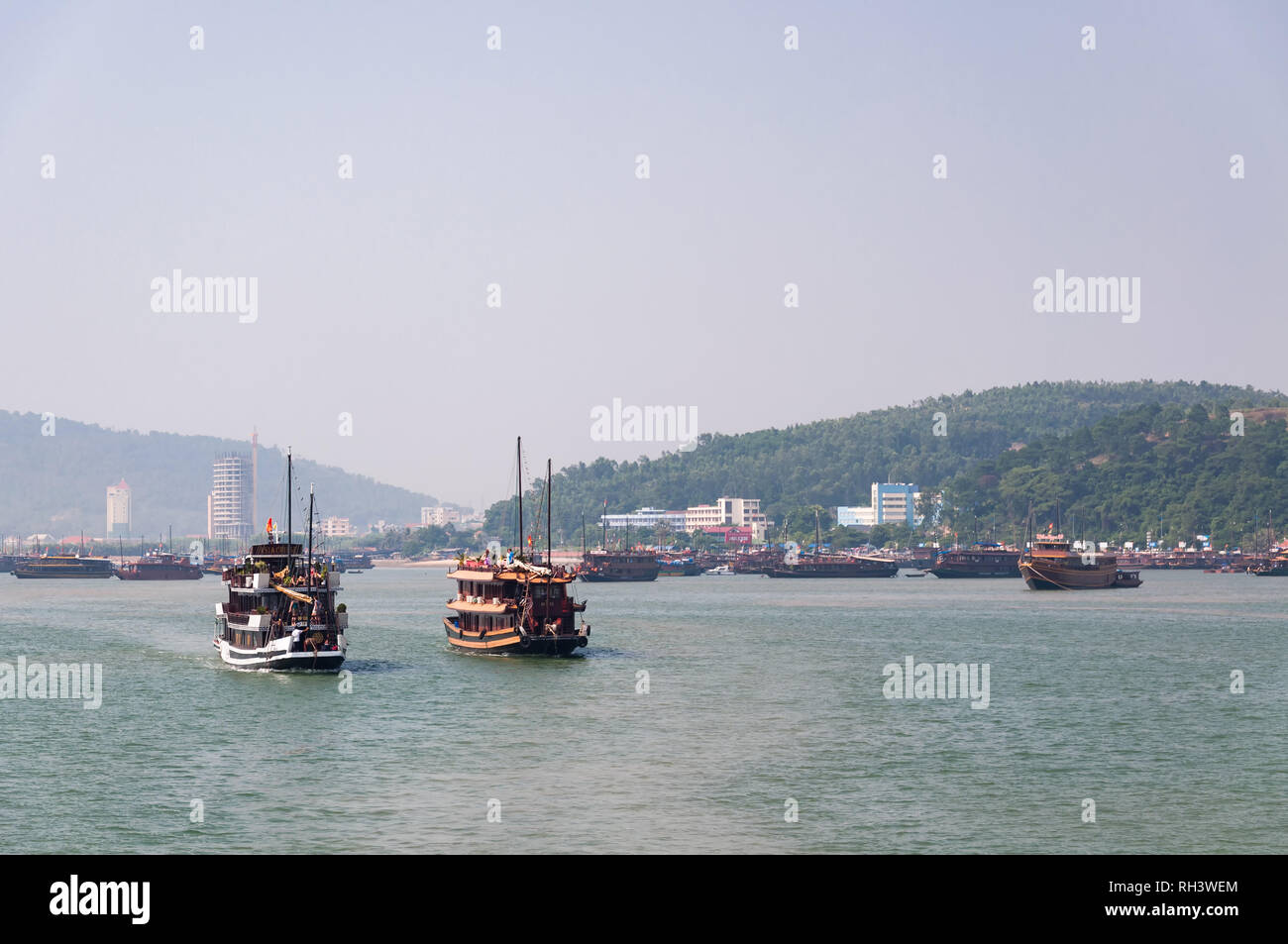 Wooden cruise ships and junks sailing from Halong or Ha Long Bay with city in background, Vietnam Stock Photo