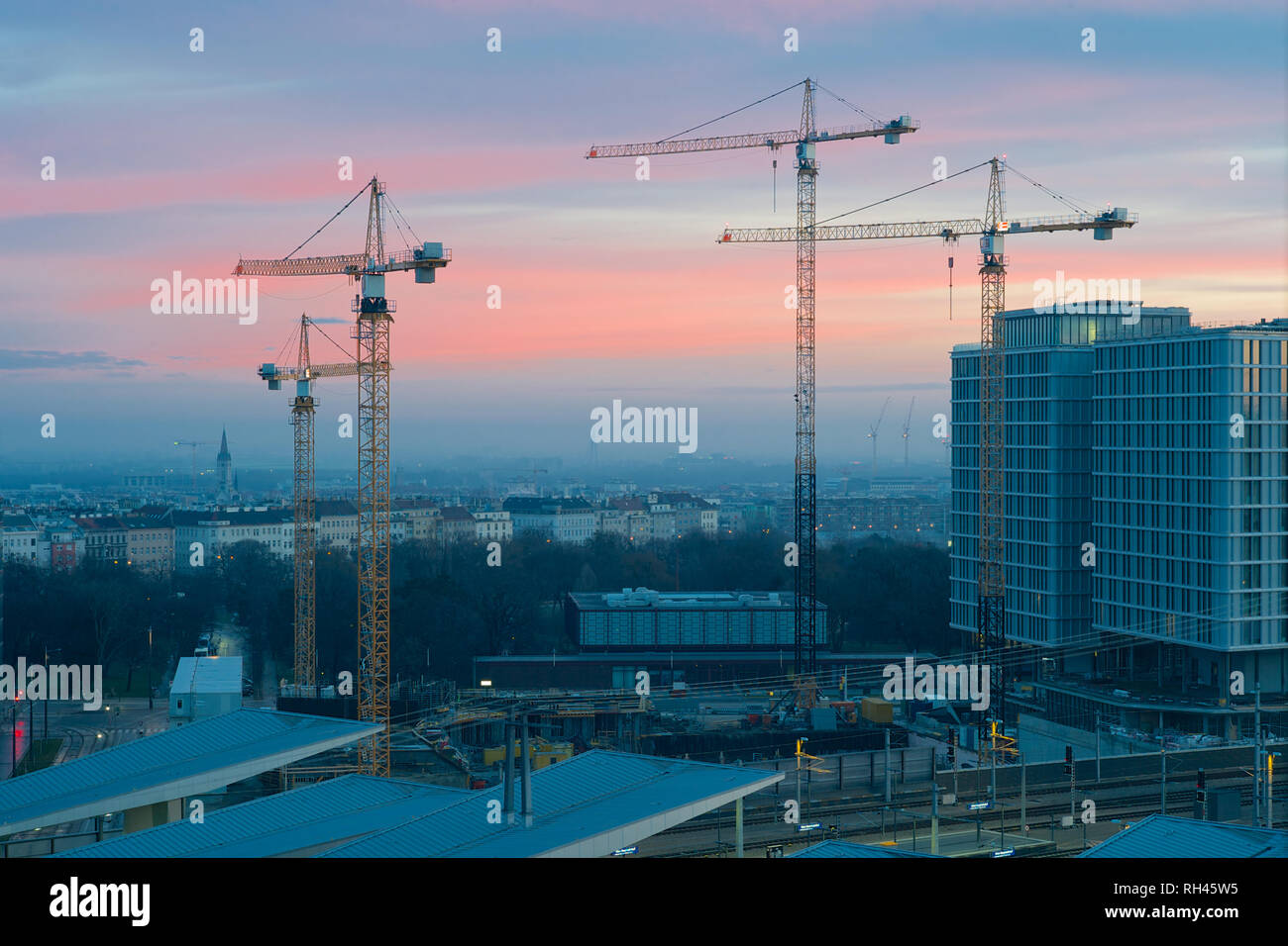 Four Construction Cranes in early morning light with the Skyline of Vienna with a dramatic sky with orange, blue and pink hues. Stock Photo