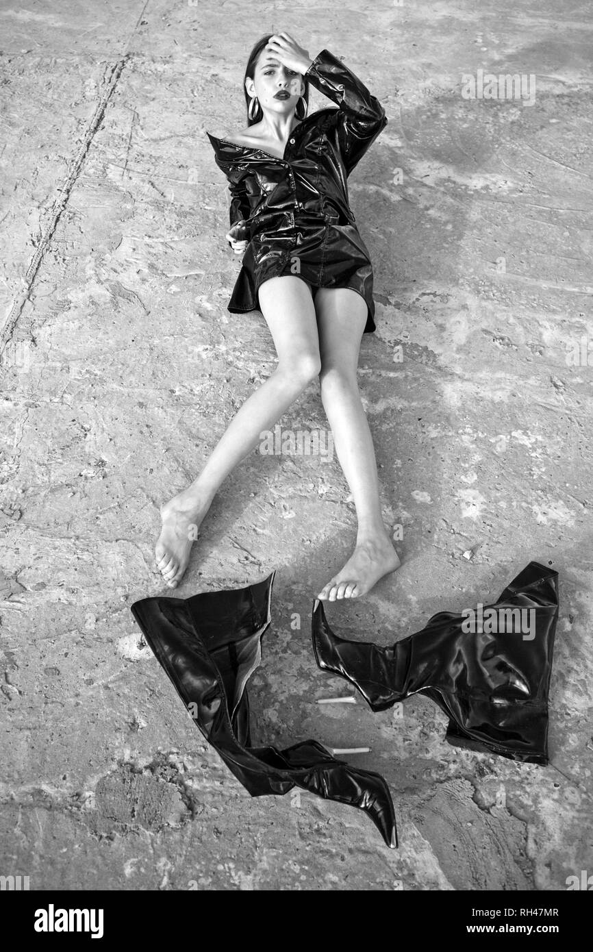 Woman in black latex jacket, barefoot without knee high boots, fashion. Girl with makeup face lie on cement ground, beauty. Fashion, style, accessory. Stock Photo