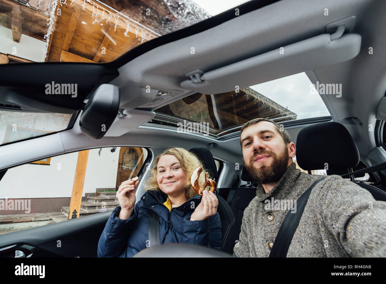 Young couple taking selfie in a car with glass roof. Wide angle lens shot. Bearded man and blonde woman smiling. Stock Photo