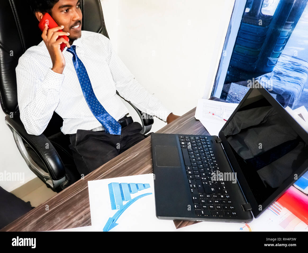 Young adult student business indian man talking with red mobile while confortable sitting in armchair in the office. Desk with graphs sheets on end pe Stock Photo