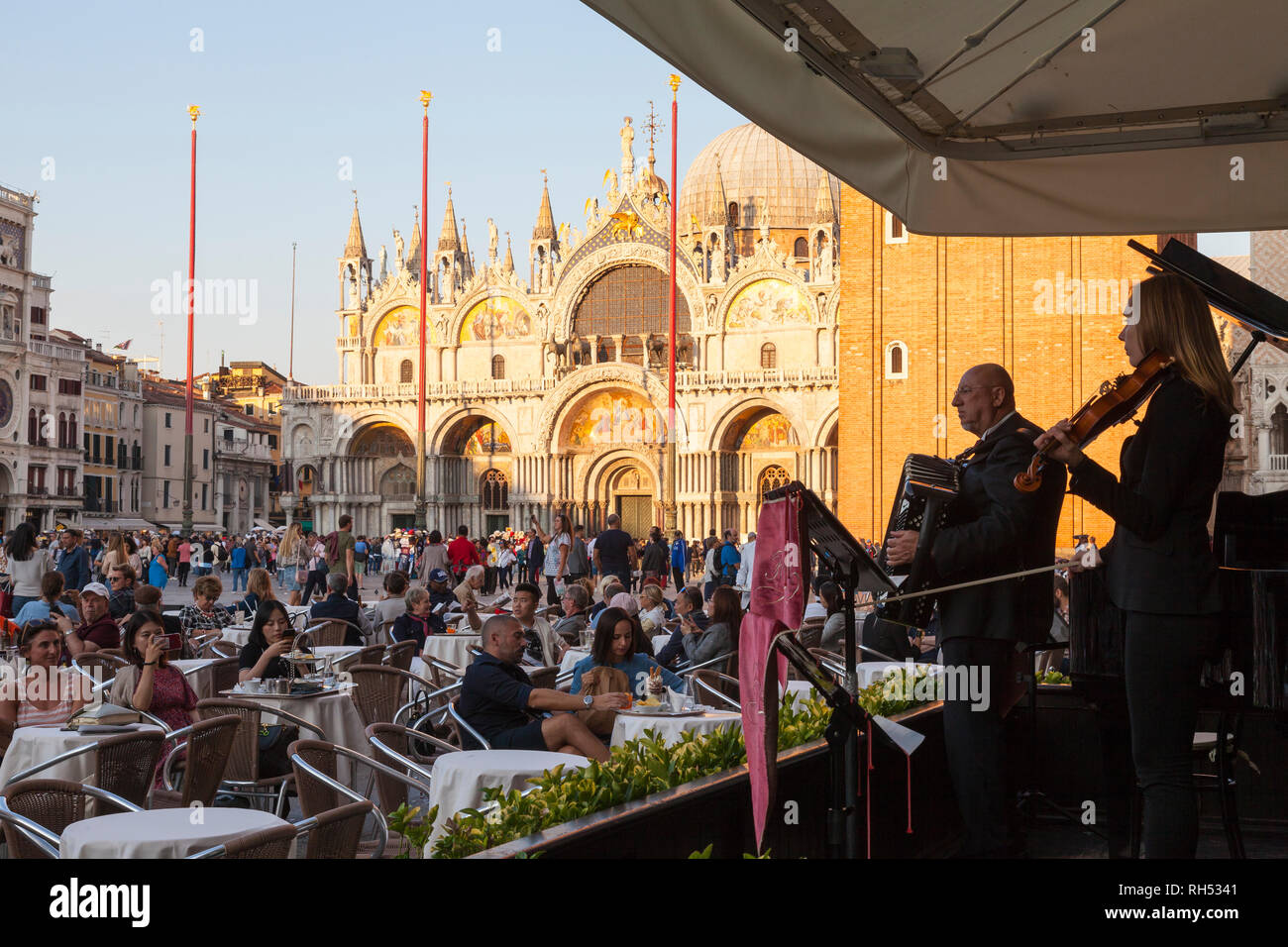 Tourists enjoying sundowners at cafe Florian in Piazza San Marco, to live music from the band  in front of St Marks Cathedral at sunset Stock Photo