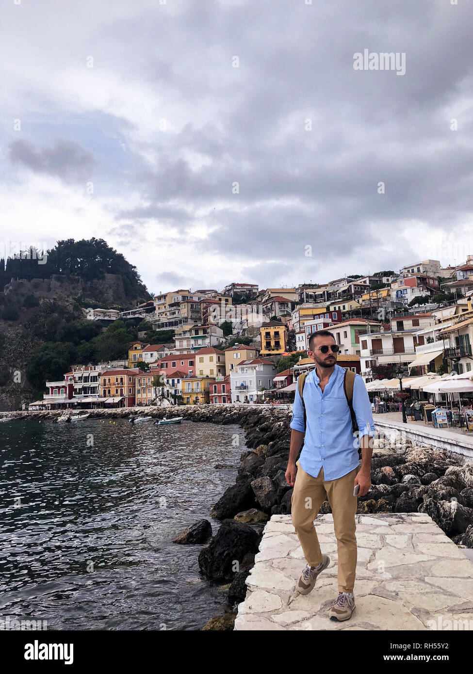 Portrait of a young man with brown pants, blue shirt, sun glasses and a brown backpack at Parga Coast Greece. Stock Photo