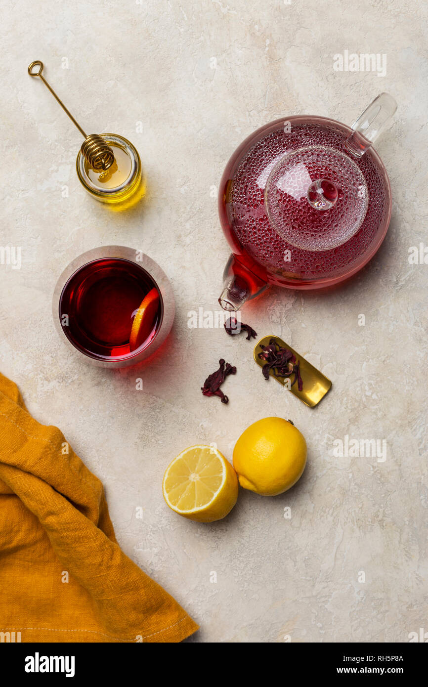 Transparency glass of hibiscus tea (karkade, Agua de flor de Jamaica) with lemon, glass teapot, honey with honey stick and orange napkin at white back Stock Photo