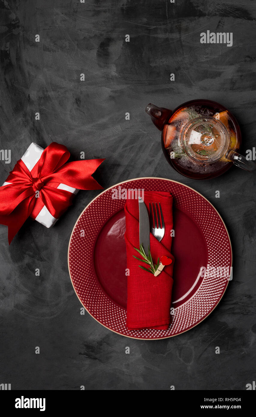 Festive serving plate for Valentine's Day with fork, knife, teapot, gift box and heart pit with rosemary brunch at plate on a black table. Top view. Stock Photo