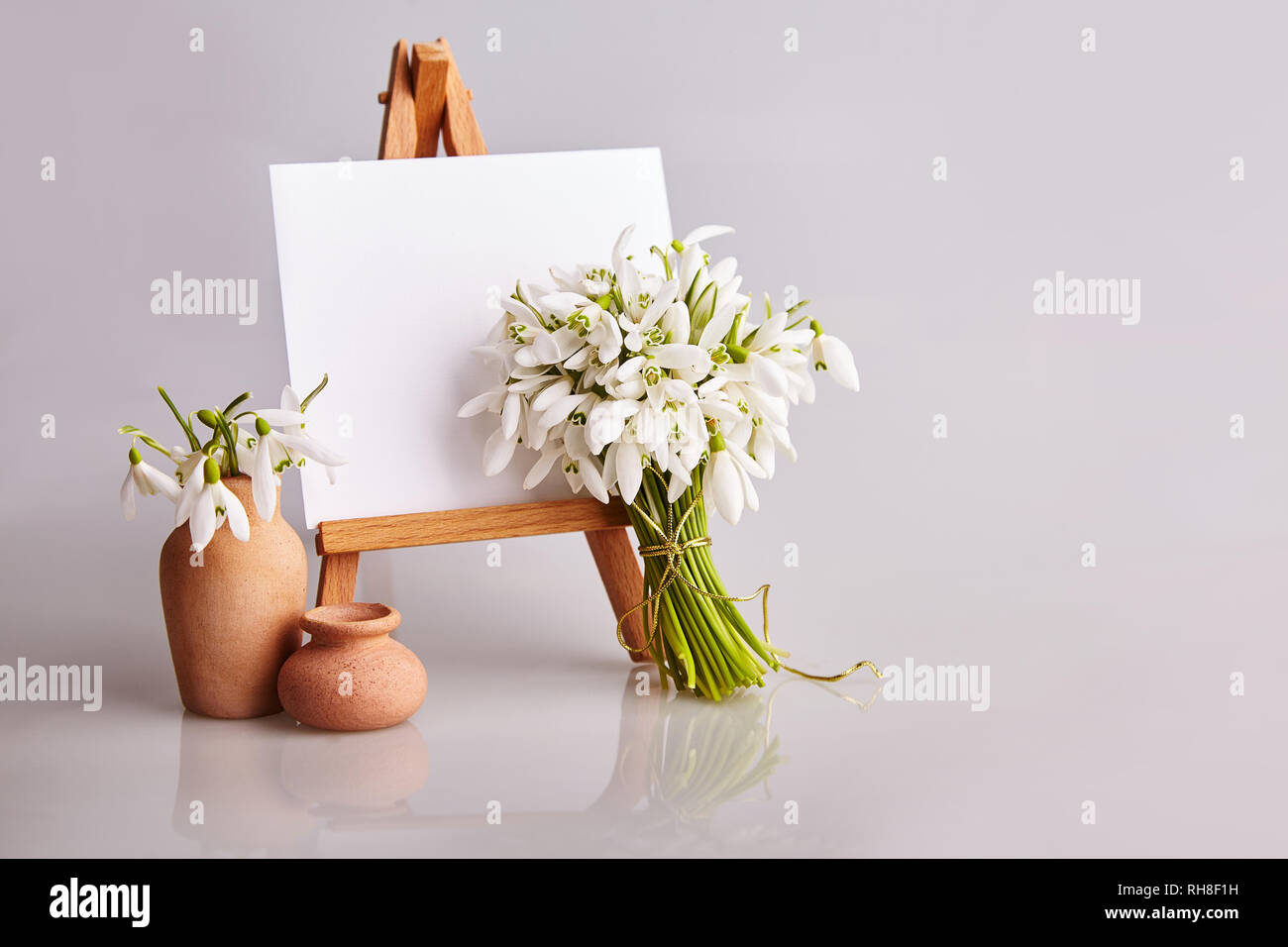 Bouquet of snowdrops and a small easel with a white paper and mini jars on a grey background. Greeting card with a space for text Stock Photo