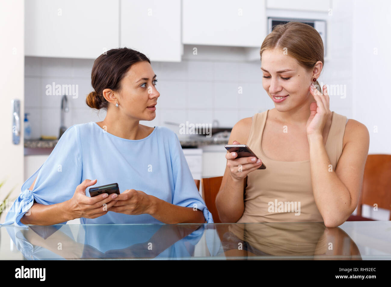 Two young women talking and using smartphone in kitchen interior Stock Photo