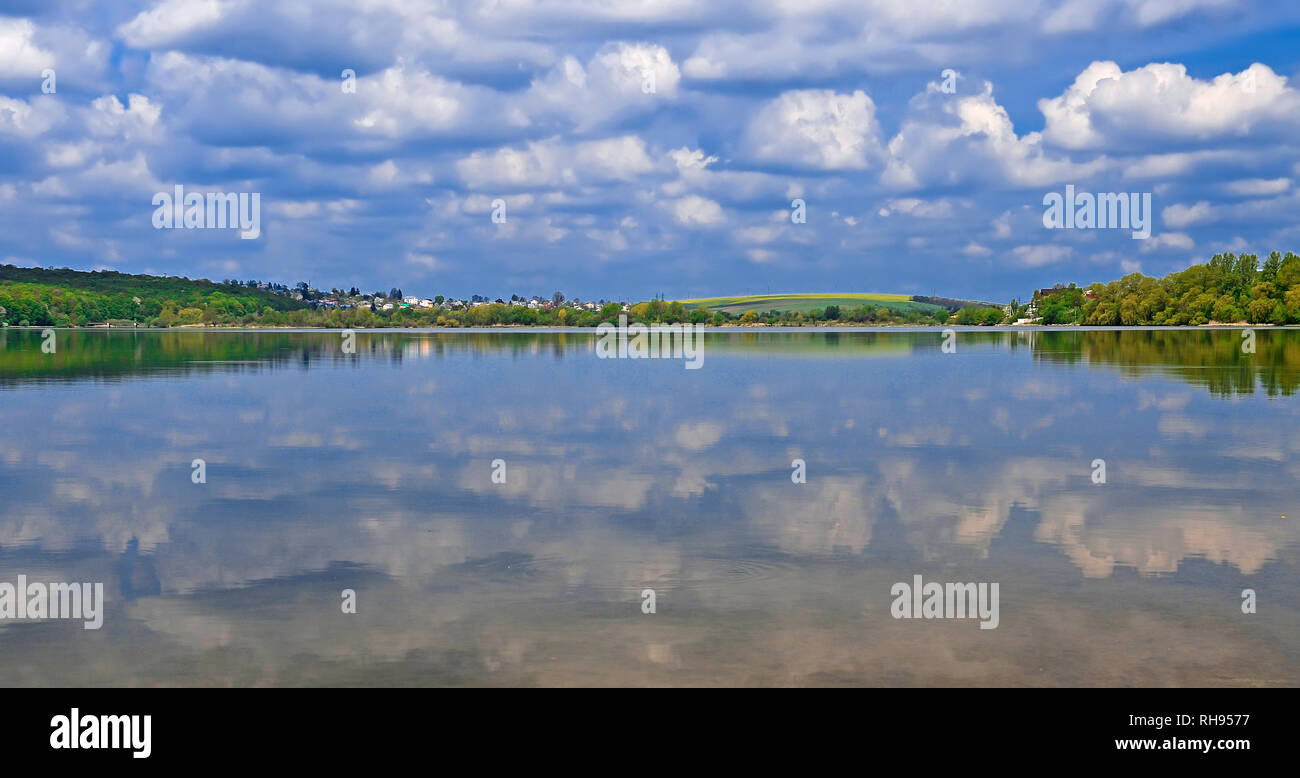 A bridge leads to an island in the middle of a lake on which tall green trees grow against a blue sky with clouds Stock Photo
