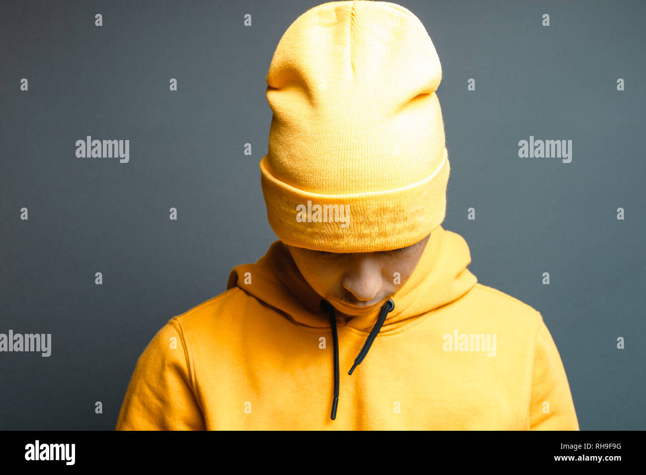 portrait of young rapper with yellow beanie cap and hoodie looking down in front of grey background Stock Photo
