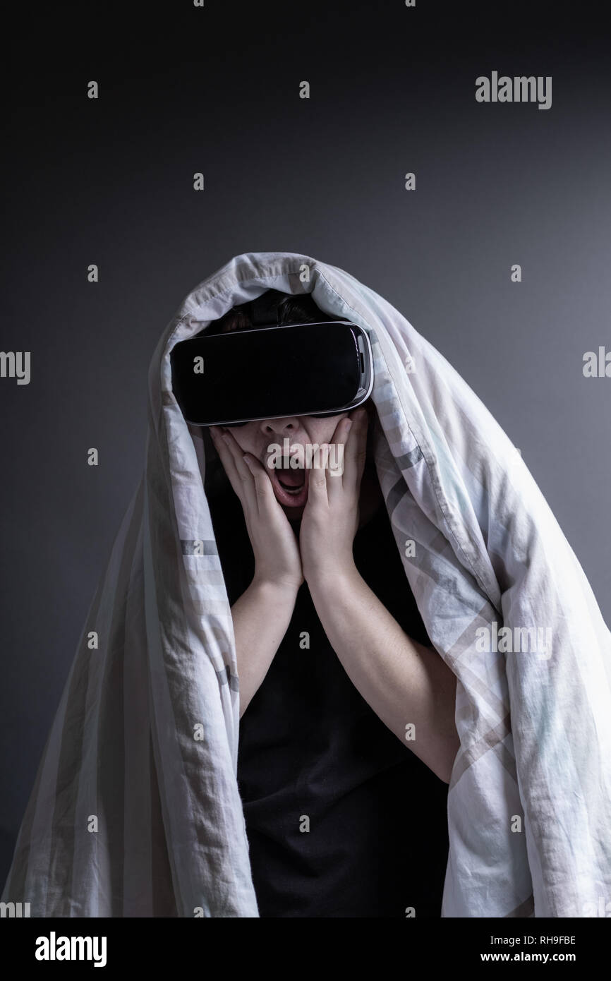 horrified young man covered up with duvet getting experience with vr device watching horror film in front of grey background. Stock Photo