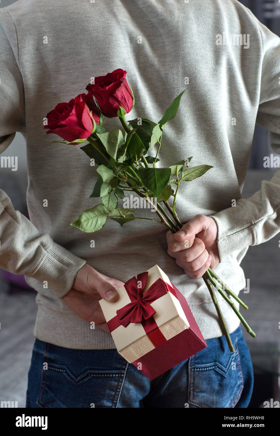 Young man holding red rose flowers and gift box behind his back at home.  Surprised gift for woman. Romantic and valentines day holiday concept Stock  Photo - Alamy