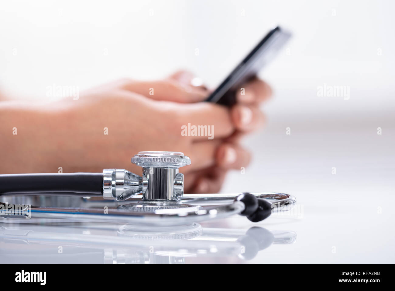 Close-up Of Stethoscope In Front Of Woman's Hand Holding Cellphone Over Reflective White Desk Stock Photo