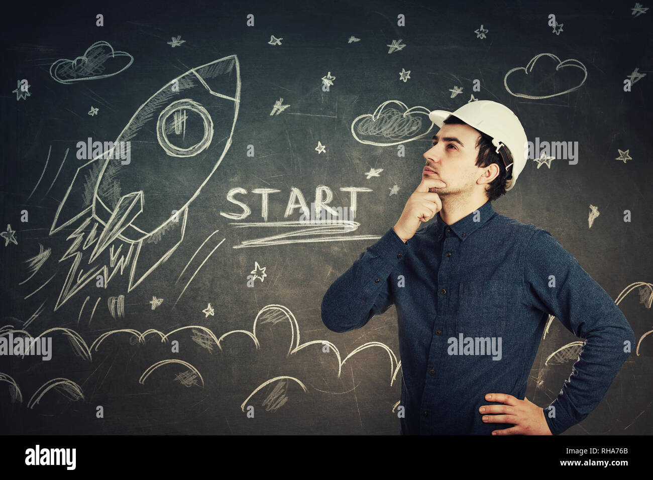 Serious thoughtful young man scientist engineer wearing protective helmet holding hand under chin pensive looking up isolated over a blackboard with r Stock Photo