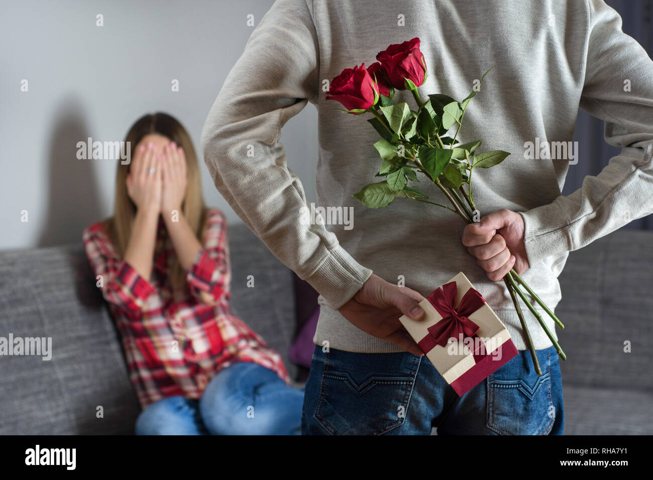 Young mans hands hiding holding chic bouquet of red roses and gift with white ribbon behind back and woman with hands over her face awaits surprise Stock Photo