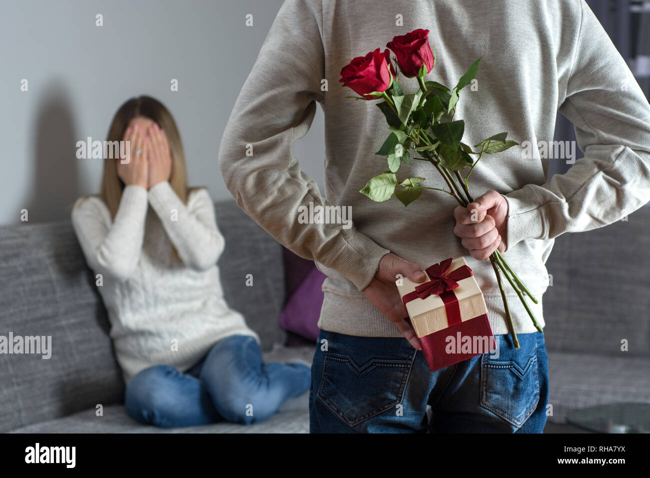 Mans hands hiding holding chic bouquet of red roses and gift with white ribbon behind back and woman with hands over her face awaits surprise in bed Stock Photo