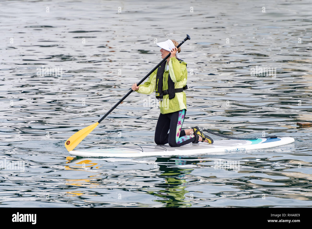 Vancouver, Canada - September 23 2017: Active caucasian adult female mid 30s kneeling on paddle board on river wearing green raincoat and visor hat Stock Photo