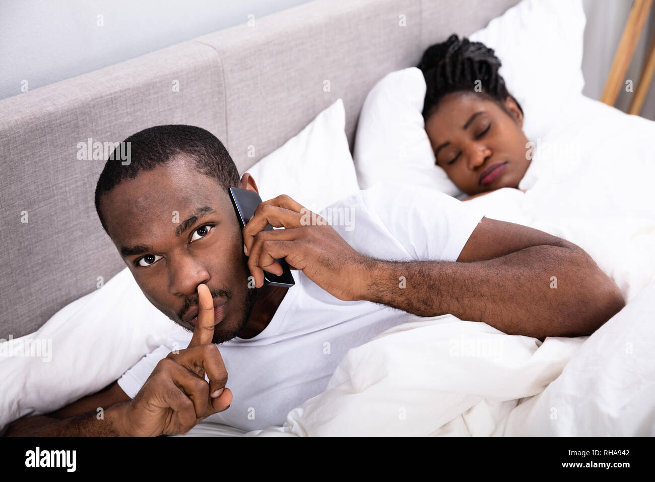 Close-up Of A Young African Man Talking Secretly On Cellphone While His Wife Sleeping Stock Photo