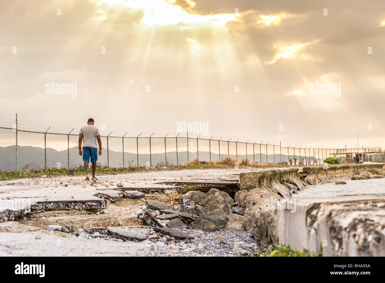 Young black male walking along road damaged by the sea, towards sun rays beaming through clouds. Concept of Hope, Broken Road, Perseverance Stock Photo