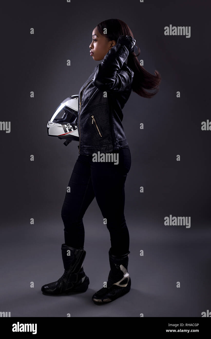 Black female motorcycle biker or race car driver or stuntwoman wearing leather racing suit and holding a protective helmet.  She is standing confident Stock Photo