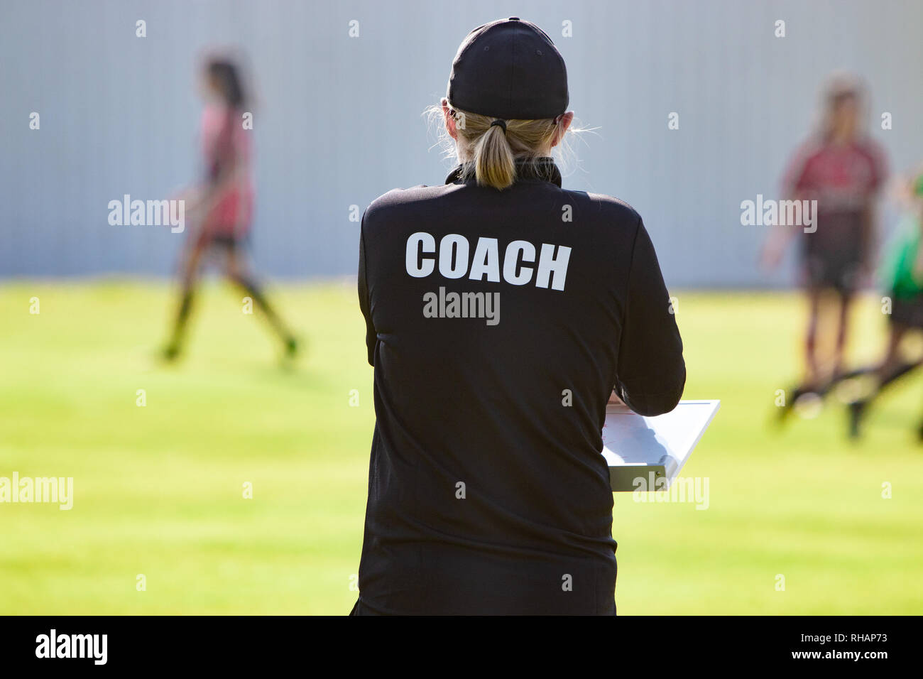 Back view of female sport coach in black COACH shirt at an outdoor sport field, watching her girl football team Stock Photo