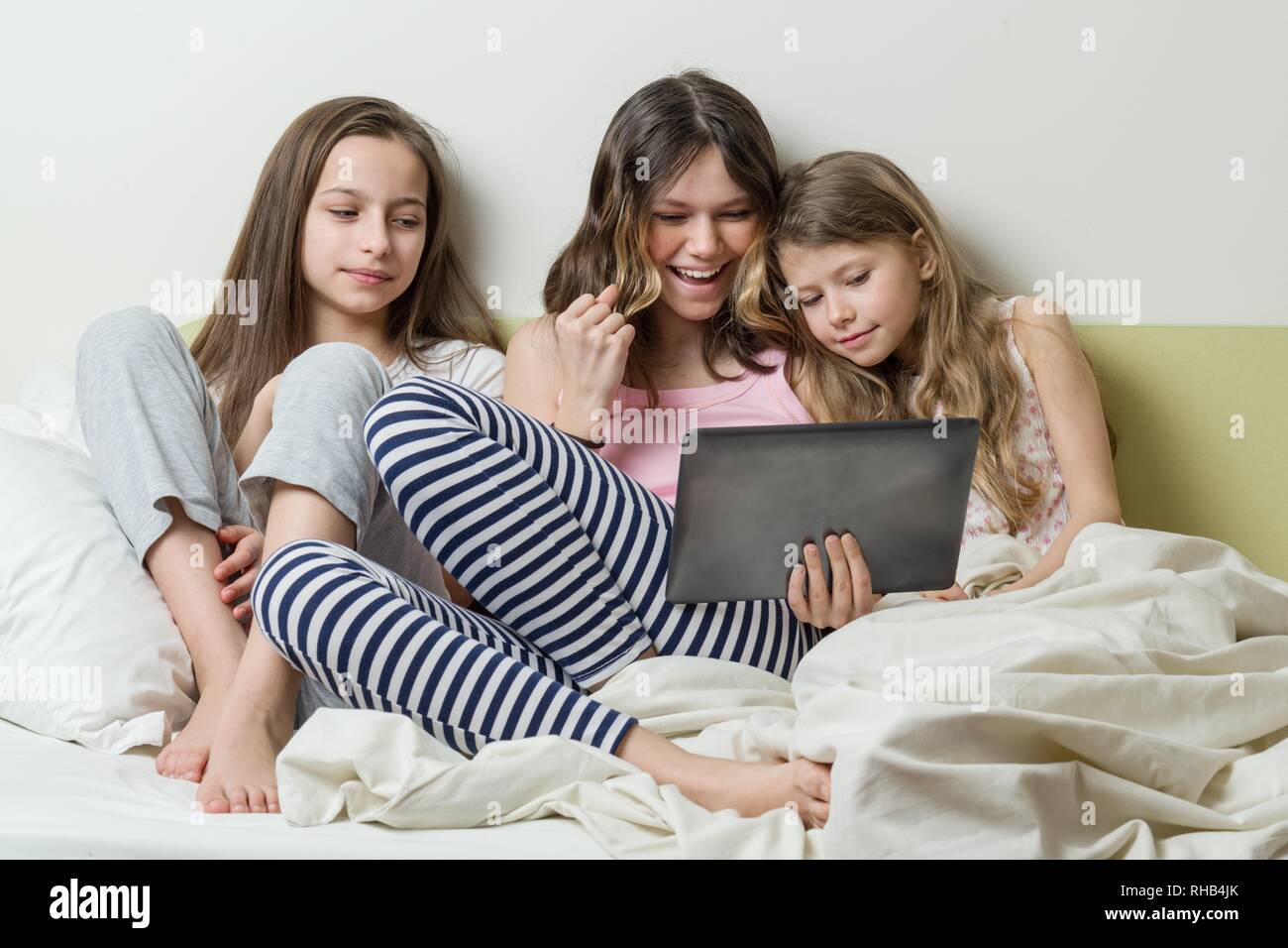 Three girls in pajamas are sitting in bed at home with interest looking ...
